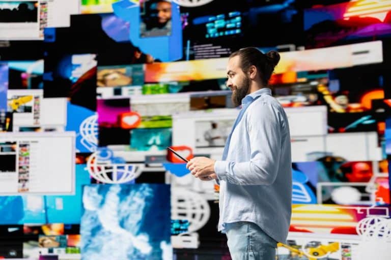 Man with a bun and beard holding a tablet in front of a vibrant wall of digital screens and graphics, wearing a light blue shirt.