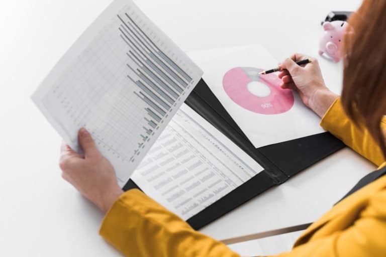 Person in a yellow jacket reviews financial charts in a binder, plotting data with a pencil beside a pink donut chart and a piggy bank nearby.