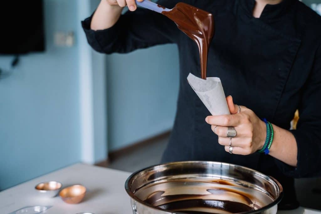 - פרסום ישראל Person in black pours melted chocolate from a spatula into a parchment cone over a bowl of chocolate.