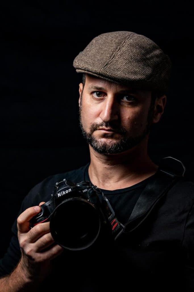 - פרסום ישראל Portrait of a man wearing a brown tweed flat cap, holding a Nikon DSLR camera against a dark background, looking at the camera.