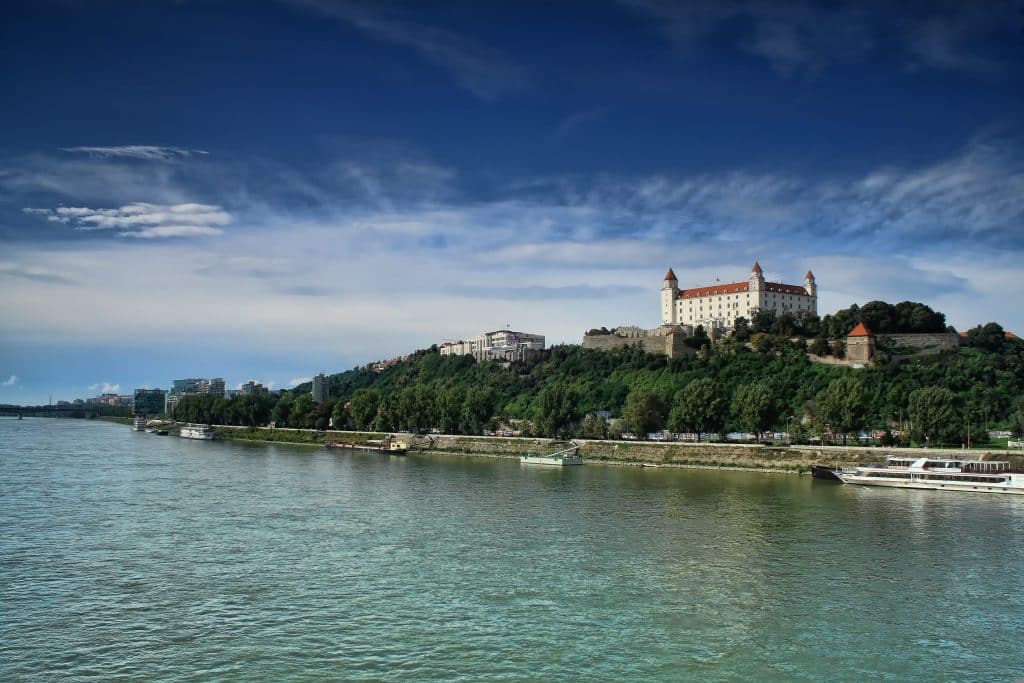 - פרסום ישראל Castle perched on a hill above a calm river, with boats along the shore and trees in the foreground under a blue sky.