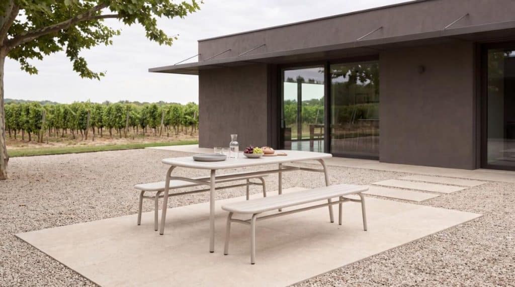- פרסום ישראל Outdoor modern patio scene: table and two benches set on a concrete pad, with plates, a carafe and fruit, beside a dark contemporary house and gravel yard; vineyard in the background.